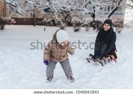 A little girl tries to ride her mom on a sled. Adults ride a sled. Winter entertainment. Mom plays with her daughter. Joke, comic picture. 