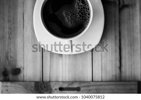 Single white cup on a wood or wooden table filled with black coffee black and white photography
