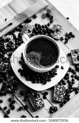 Single white cup on a wood or wooden table filled with black coffee beans and almond chocolate bars black and white photography
