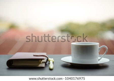 Cup of coffee and notebook with pen on table and natural view background and texting space. Outside view of coffee cafe for relaxation, read book, write note, drink coffee in free time. Still life.