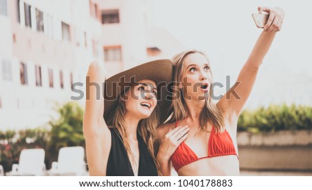 Two beautiful girls having fun taking selfie photo with smart phone camera at swimming pool