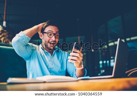 Pleasantly surprised young male in stylish eyeglasses received sms message with cash prize on smartphone device.Happy man watching winning football match results on internet website. Excited news Royalty-Free Stock Photo #1040364559