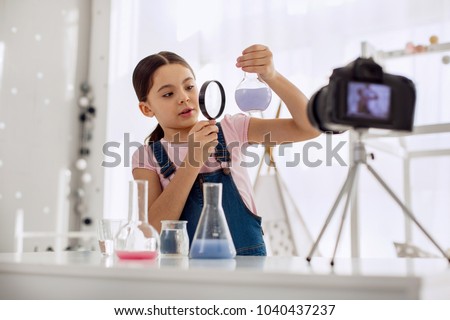 Talented chemist. Lovely pre-teen girl looking at a flask with a chemical in it through a magnifying glass while recording herself for a video blog Royalty-Free Stock Photo #1040437237
