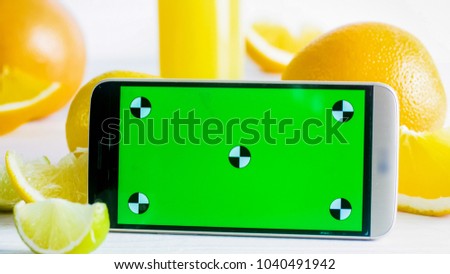 Closeup image of mobile phone with green chromakey screen next to fresh fruits on white table. Perfect for inserting your own photo