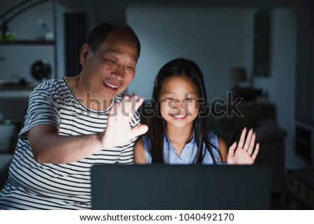 Little girl and her grandfather are using a laptop at home to video call their family. It is dark and the screen is illuminating their faces while they wave to the screen. 