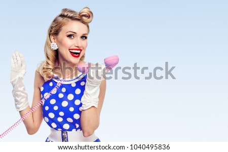 Funny portrait of beautiful happy woman with phone, dressed in pin-up style dress in polka dot and white gloves, with copyspace area for slogan or advertising text message, on blue background.
