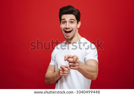 Portrait of a smiling young man in white t-shirt pointing finger at camera isolated over red background