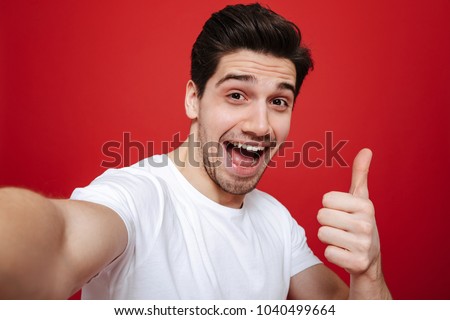 Portrait of a happy young man in white t-shirt showing thumbs up gesture while taking a selfie isolated over red background