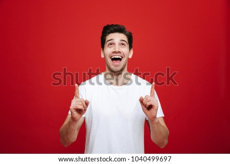 Portrait of a joyful young man in white t-shirt pointing fingers up at copy space isolated over red background