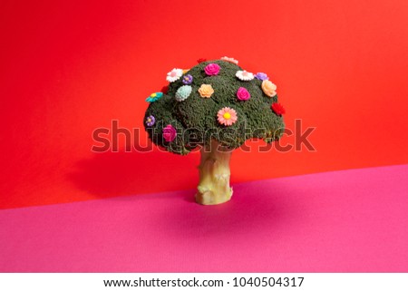 a diversion of a standing broccoli covered with numerous plastic flowers isolated on a vibrant pink and red background. Humorous metaphor of a bouquet of flowers. Minimal color still life photography