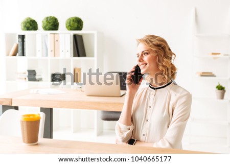 Photo of pretty young business woman in office looking aside talking by mobile phone.