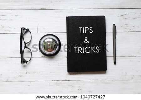 Top view of eyeglasses, ring bell,pen and notebook written with TIPS & TRICKS on white wooden background.