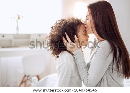 Sweet kisses. Beautiful happy young long-haired mother smiling and kissing her daughter and hugging her