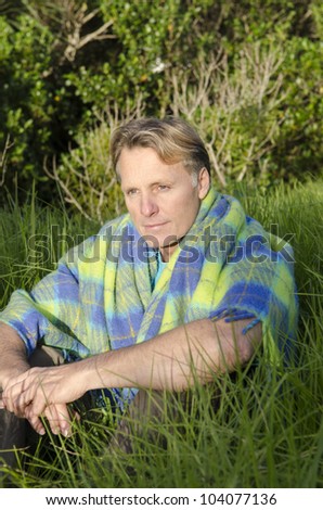 A man sitting in the grass wrapped in a green and blue colored blanket.