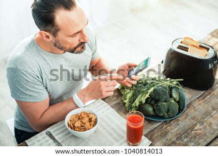 Is under control. Concentrated careful unshaken man sitting in the kitchen by the table using his cellphone preparing for eating. Royalty-Free Stock Photo #1040814013