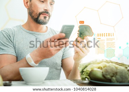 Vitamins and energy. Pleasant satisfied unshaken man using cellphone holding and overlooking a broccoli. Royalty-Free Stock Photo #1040814028