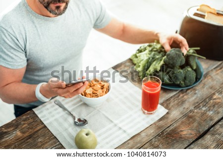 Program of nutrition. Occupied concentrated unshaken man having breakfast by the table looking at smartphone taking vegetables. Royalty-Free Stock Photo #1040814073