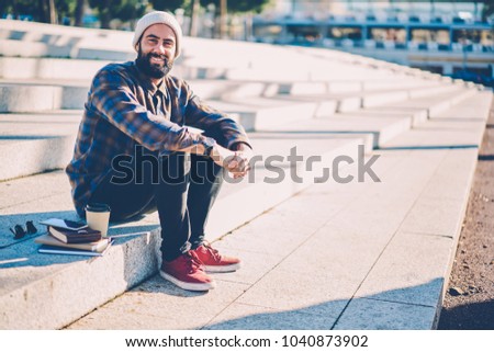 Portrait of cheerful bearded hipster guy dressed in stylish casual clothes smiling at camera while sitting on stairs in urban setting with notepads and coffe to go enjoying leisure time outdoors