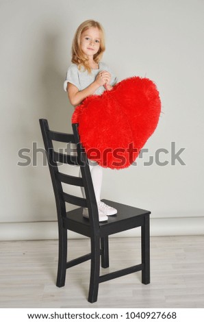 A little cute blonde girl stands on a chair and holds a heart