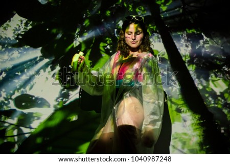 The picture of a deep wild forest is projected on a young woman in a raincoat with banana in her hand.