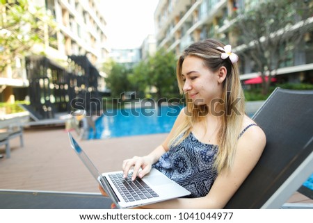 young woman female working uses internet and laptop outdoors remotely as freelancer close to blue swimming pool and apartment building on sunny day with sunshine. Concept of working in