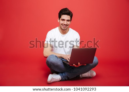 Portrait of a happy young man in white t-shirt holding laptop computer while sitting on a floor isolated over red background