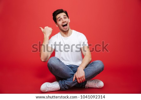 Portrait of a joyful young man in white t-shirt sitting on a floor and pointing finger away isolated over red background