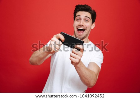Portrait of an excited young man in white t-shirt playing games on mobile phone isolated over red background