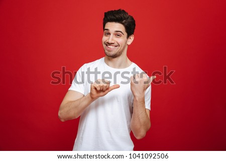 Portrait of a happy young man in white t-shirt pointing fingers away at copy space isolated over red background
