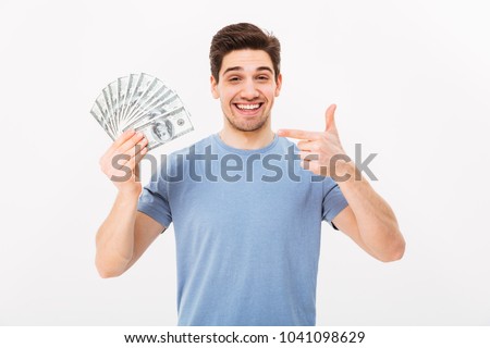 Photo of happy winner man in casual t-shirt smiling and pointing finger on fan of money in dollar banknotes isolated over white wall