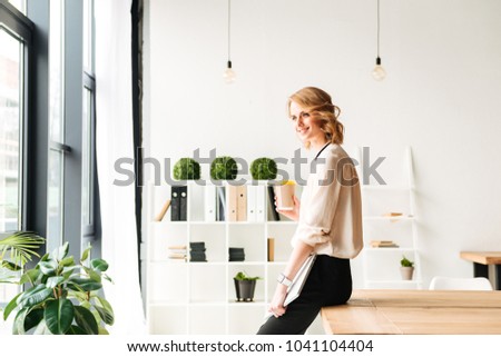 Photo of cheerful young business woman in office holding laptop computer looking aside drinking coffee.
