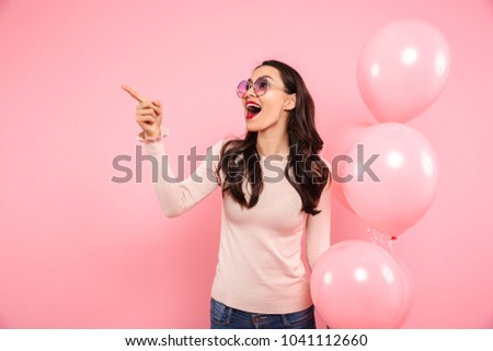 Photo of delighted adult girl with long dark hair in round glasses holding lots of balloons and pointing finger aside isolated over pink background