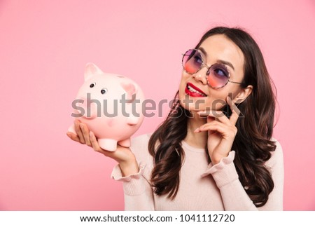 Smiling successful woman 20s with red lips in trendy glasses holding piggy bank on palm isolated over pink background