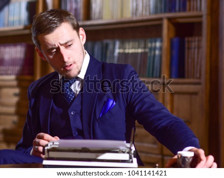 Writer or author with typewriter looks for inspiration for new book, drinks coffee in library. Retro writer concept. Man in suit with busy face in library with antique books on background, defocused.