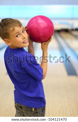 Smiling boy dressed in blue T-shirt holds pink ball in bowling club, stands with his back to camera and turns; shallow depth of field