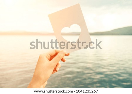 Card with a carved heart in a woman's hand against a background of blue mountains and water with clouds. Ocean, sea, lake.
