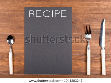 Black paper with the word Recipe written next to cutlery on brown wooden table. Mockup.
