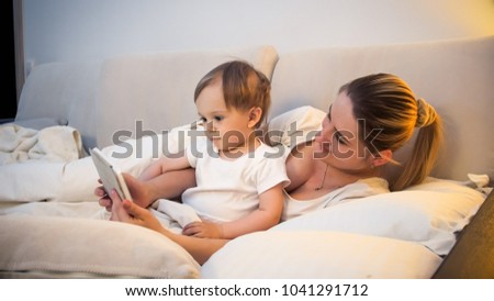 Portrait of cute toddler boy lying with mother in bed and watching cartoons