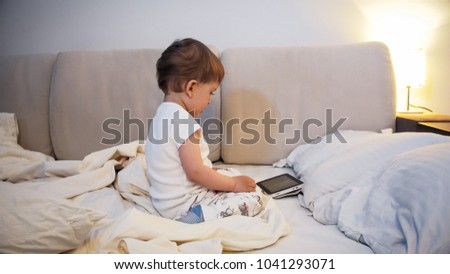 Toddler boy in pajamas sitting o nbed at night and watching cartoons on digital tablet