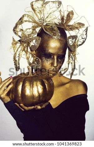 halloween golden woman or girl holding painted gold pumpkin has pretty face with makeup and body art metallized color with decorative flowers on head on white grey background