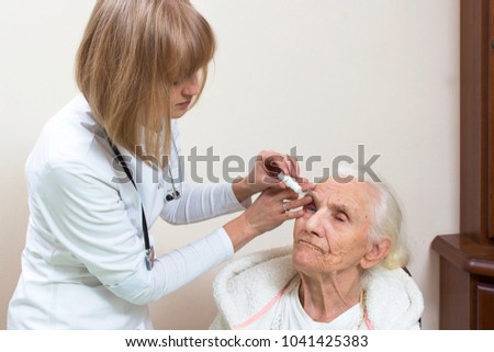 The doctor lets drops into the eye of a very old woman sitting on a chair in a white bathrobe. The doctor's woman's hands let drops into the eye of a very old wrinkled woman sitting on a chair
