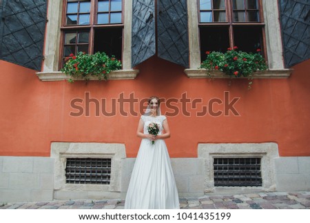 Beautiful bride in lace satin dress with pink rose bouquet is standing near the orange ancient stone walls. Old town windows with shutters on background. Elegant wedding photo portrait.