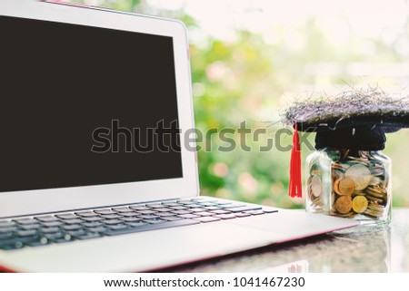 Computer laptop and square academic cap with the glass jar of coin against blurred natural green background for education, finance and saving money concept