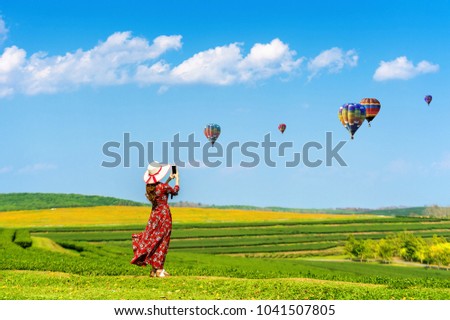 Woman standing on green grass and taking a photo to hot air balloons with smartphone.