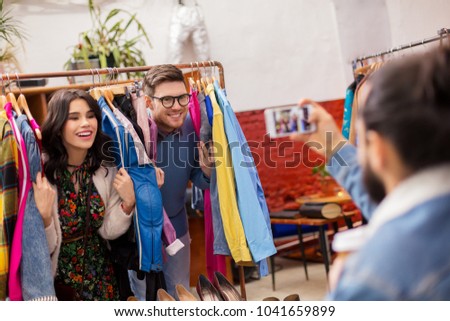 sale, shopping, fashion and people concept - friend photographing happy couple having fun by smartphone at vintage clothing store hanger