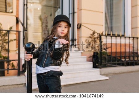 Outdoor portrait of cute little girl in hat sending air kiss with surprised face expression. Amazed long-haired girlie in rock style outfit gladly posing on the street enjoying good day