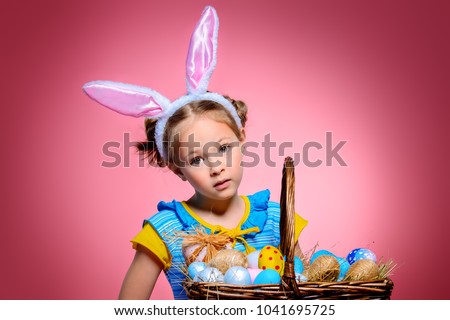 Easter holiday. Portrait of a happy little girl wearing bunny ears with basket full of painted eggs over pink background.