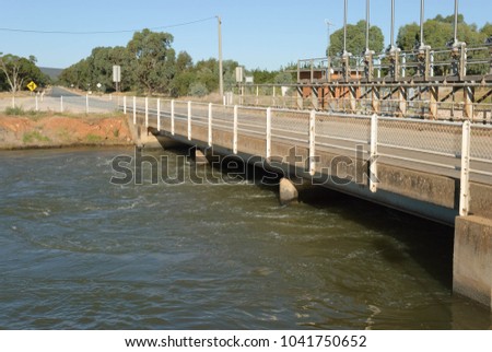 an overpass over an irrigation channel with water flowing underneath and a building and trees in the background and a blue cloudless sky