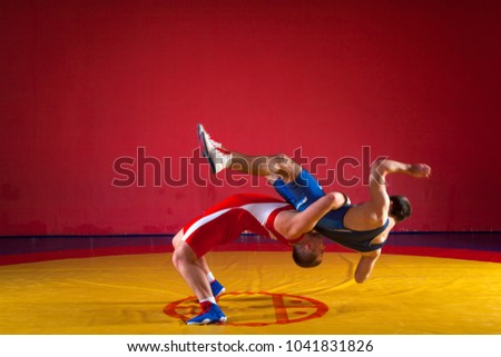 Two young men wrestlers in blue and red wrestling tights are wrestlng and making a hip throw on a yellow wrestling carpet in the gym, sied view