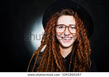 Studio shoot of girl in black with dreads, hat and glasses at black background.